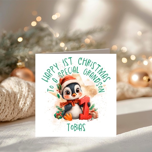 Christmas card with a penguin and text 'Happy 1st Christmas to a special grandson Tobias' on a blurred festive background.