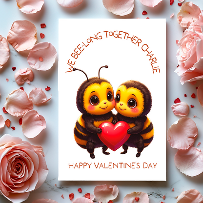 Valentine's Day card with two bees holding a heart and the text 'We bee-long together' and Happy valentines Day' surrounded by flowers and petals.
